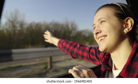 Free Girl Travels By Car Catches Wind With Her Hand From Car Window. Young Woman With Long Hair Sits In Back Seat Of Car, Reaches Out To Window And Catches Glare Of Sun. A Teenager Travels In A Car.