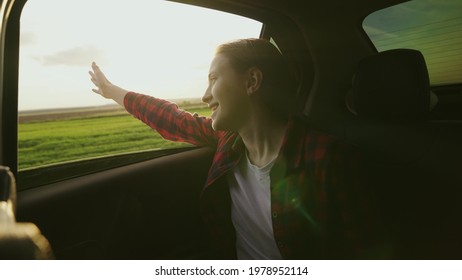 Free Girl Travels By Car Catches Wind With Her Hand From Car Window. Young Woman With Long Hair Sits In Back Seat Of Car, Reaches Out To Window And Catches Glare Of Sun. A Teenager Travels In A Car.