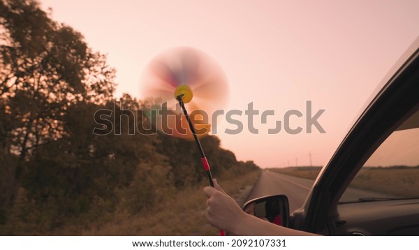 free girl waving a pinwheel out of the car window\
in wind, travel adventure road at pink sunset, family car ride,\
reach out of car window in sunlight of sky, enjoy movement,\
childhood dream concept