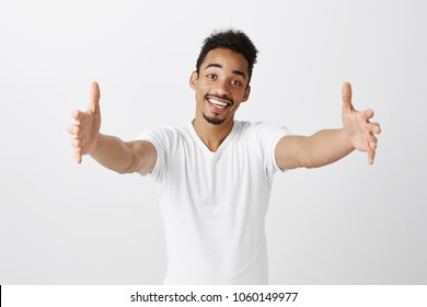 Free hugs for person who had bad day. Cheerfull kind dark-skinned male model in white t-shirt, pulling hands towards camera, wanting to cuddle, smiling broadly, welcoming friend to come into his arms