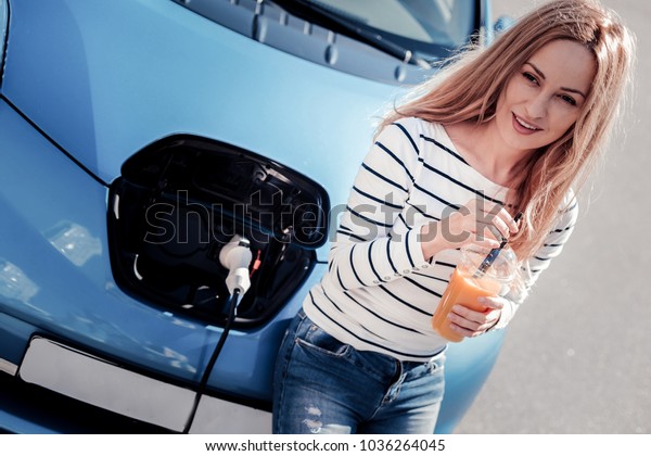 Free minute. Calm pretty pleasant woman drinking\
juice near her car looking aside and waiting when the car will be\
charge.