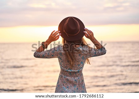 Free serene spirit boho woman wearing dress and felt hat standing back by the seashore at sunset and enjoying calm and freedom