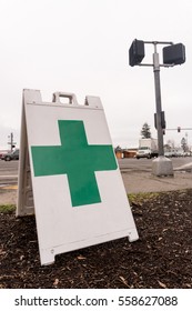 A Free Standing Sign With A Green Cross Shows That A Marijuana Dispensary Is Nearby High Traffic Busy Intersection.