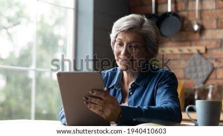 Free time with gadget. Positive elderly hispanic granny relax at home by window use tablet device contact children online chat in app read latest news. Happy mature woman surf internet on digital pad