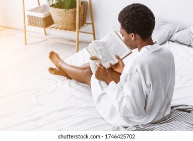 Free time to read. African american girl with cup of coffee passionate about book on bed, sun flare, free space