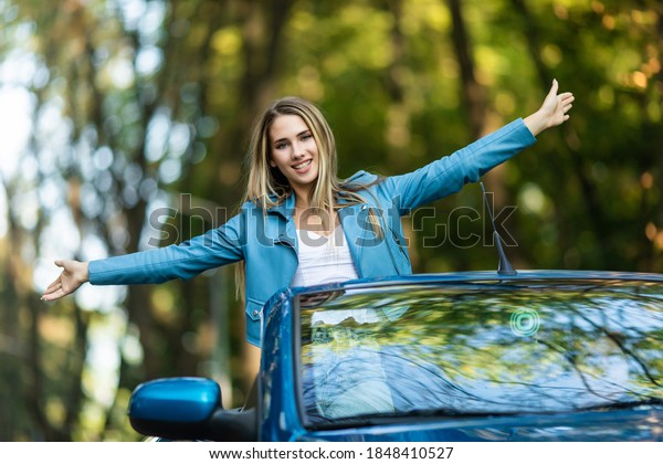 Free woman in cabriolet\
cheering joyful with arms raised. Woman going on road trip travel\
on summer day