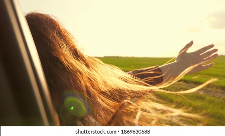 Free Woman Travels By Car Catches The Wind With Her Hand From The Car Window. Girl With Long Hair Is Sitting In Front Seat Of Car, Stretching Her Arm Out Window And Catching Glare Of Setting Sun