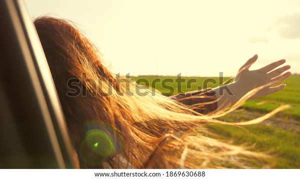 Free woman travels by car catches the wind with her\
hand from the car window. Girl with long hair is sitting in front\
seat of car, stretching her arm out window and catching glare of\
setting sun