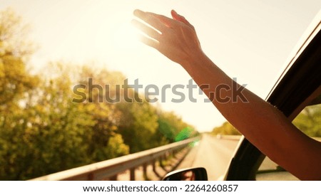 Free young girl waves her hand from car window, travels, catches sun glare with her fingers. Auto travel on road on vacation. Young woman driver plays catches fresh wind with hand from car window.