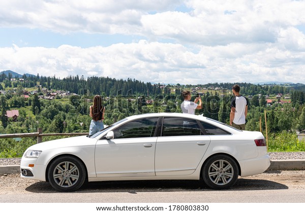 Freedom of car travel. Car with three friends at\
the top of the hill with beautiful view on mountains, people making\
selfie using smart\
phones