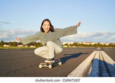 Freedom and happiness. Happy asian girl riding her longboard on an empty sunny road, laughing and smiling, skateboarding.