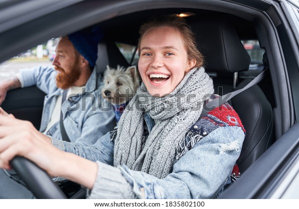 Freedom of the open road. Side\
view of joyful young man relaxing on the front seat while her\
girlfriend sitting near, driving their car and laughing at the\
camera