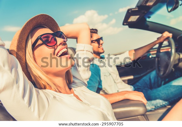 Freedom of the open road. Side view of\
joyful young woman relaxing on the front seat while her boyfriend\
sitting near and driving their convertible\

