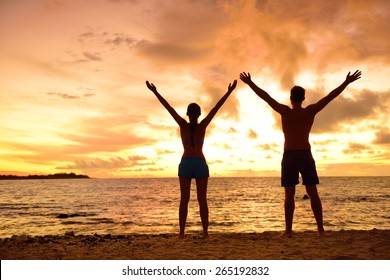 Freedom People Living A Free, Happy, Carefree Life At Beach. Silhouettes Of A Couple At Sunset Arms Raised Up Showing Happiness And A Healthy Lifestyle Against A Colorful Sky Of Clouds Background.