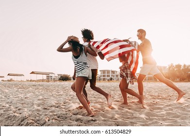 Freedom is in their veins. Cheerful young people carrying American flag while running along the beach together - Powered by Shutterstock