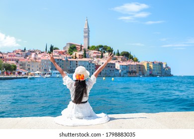 Freedom tourist woman holding her hands up with two finger making peace sign sitting on Harbor and looking to beautiful rovinj city. Young Asian lady traveling in the summer in Croatia, Europe.