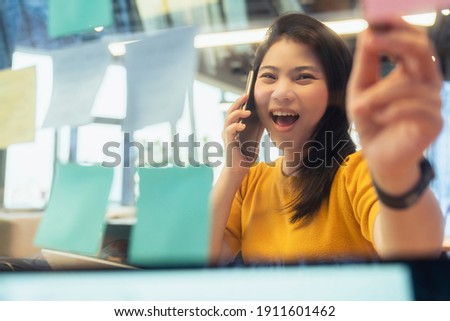 freelance asian female creative person wear casual yellow cloth conversation with smartphone hand point paper note reminder at coworking space new lifestyle with thoughful and freshness emotion