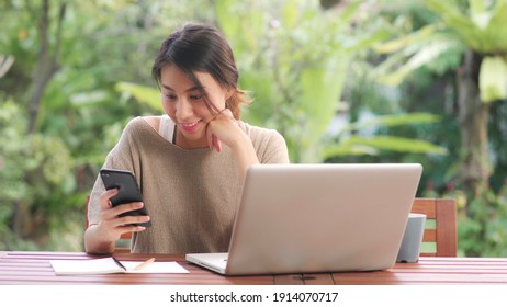 Freelance Asian Woman Working At Home, Business Female Working On Laptop And Using Mobile Phone Drinking Coffee Sitting On Table In The Garden In Morning. Lifestyle Women Working At Home Concept.