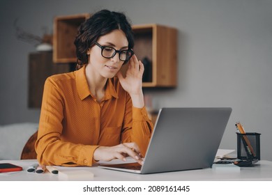 Freelance. Beautiful Focused Spanish Woman In Glasses Sits At Desk In Living Room Types Communicates With Client Live On Laptop, Focused Young Female Consultant Adviser Working Remotely Online At Home