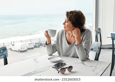 Freelance business woman enjoying a fragrant coffee alone in cafe in the morning near panoramic seaside window. - Powered by Shutterstock