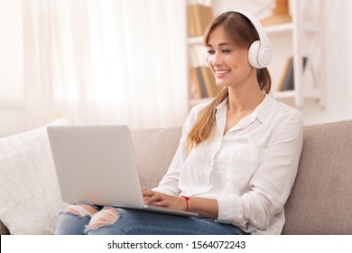 Freelance Career. Cheerful Lady In Wireless Headphones Working On Laptop Computer Sitting On Sofa At Home. Selective Focus