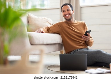 Freelance Career. Portrait Of Excited Middle Eastern Male Holding Cell Phone Working On Pc At Home Office, Happy Guy Using Computer Sitting On Floor Leaning On Sofa In Living Room, Looking Away