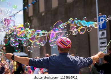 A Freelance Clown Blowing Hundreds Of Tiny, Small And Big Bubbles At Outdoor Festival In City Center. Concept Of Entertainment, Birthdays. Kids Having Fun. Shower Of Bubbles Flying In The Happy Crowd
