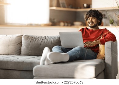 Freelance Concept. Handsome Young Indian Man Using Laptop While Sitting On Couch At Home, Millennial Eastern Guy Working Remotely On Computer In Living Room, Enjoying Online Work, Copy Space - Powered by Shutterstock