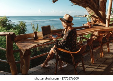 Freelance concept. Pretty young woman using laptop in cafe on tropical beach in outdoor cafe terrace with sea view. Work and travel - Shutterstock ID 1770576509