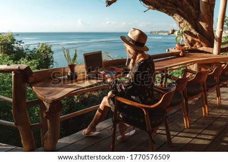 Freelance concept. Pretty young woman using laptop in cafe on tropical beach in outdoor cafe terrace with sea view. Work and travel