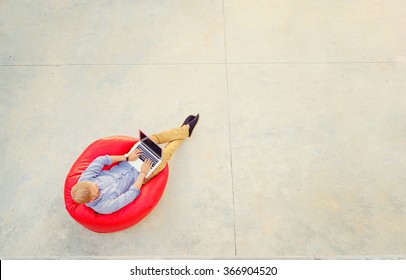 Freelance Concept. Top View Of Young Man Working On Laptop Sitting On The Red Bean Bag.