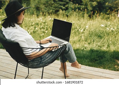 Freelance. Hipster girl with laptop sitting on wooden porch with beautiful view on woods and mountains. Stylish Young woman in hat using laptop, shopping or working online outside. - Powered by Shutterstock