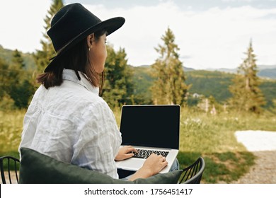 Freelance. Hipster girl with laptop sitting on wooden porch with beautiful view on woods and mountains. Stylish Young woman in hat using laptop, shopping or working online outside. - Powered by Shutterstock