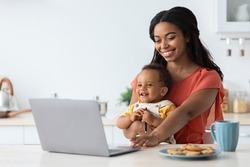 Freelance And Motherhood Concept. Black Young Woman Working Remotely On Laptop At Home And Taking Care About Baby, African American Mom Holding Infant Son And Using PC Computer In Kitchen, Copy Space