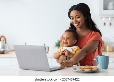 Freelance And Motherhood Concept. Black Young Woman Working Remotely On Laptop At Home And Taking Care About Baby, African American Mom Holding Infant Son And Using PC Computer In Kitchen, Copy Space