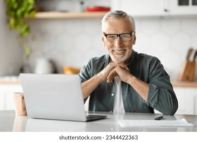 Freelance In Older Age. Happy Senior Man Sitting At Desk With Laptop In Kitchen Interior, Handsome Elderly Gentleman Using Computer For Online Work From Home, Smiling And Looking At Camera - Powered by Shutterstock