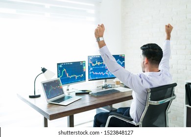 Freelance stock broker celebrating successful trading while sitting with arms raised and working at home