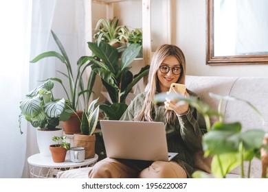 Freelance Woman In Glasses With Mobile Phone Typing At Laptop And Working From Home Office. Happy Girl Sitting On Couch In Living Room With Plants. Distance Learning Online Education And Work.