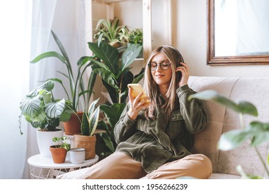 Freelance woman in glasses with mobile phone listening music in headphones and relax at home. Happy girl with closed eye sitting on couch in living room with plants. 