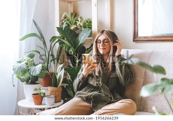 Freelance woman in glasses with\
mobile phone listening music in headphones and relax at home. Happy\
girl with closed eye sitting on couch in living room with plants.\
