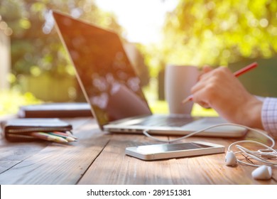 Freelance work.
Casual dressed man sitting at wooden desk inside garden working on computer pointing with color pen electronic gadgets dropped around on table side view