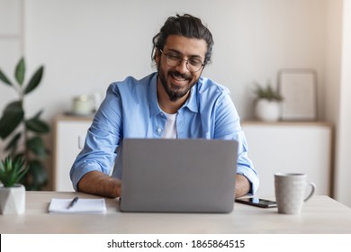 Freelance Work. Happy Millennial Indian Man Working On Computer At Home Office, Sitting At Desk With Laptop, Handsome Western Guy Looking At Device Screen And Smiling, Enjoying Remote Job, Copy Space