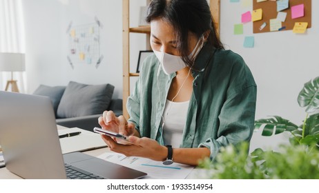 Freelancer Asia women wear face mask using smartphone shopping online via website while sitting at desk in living room. Working from home, remotely work, social distancing, quarantine for coronavirus. - Powered by Shutterstock