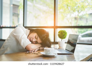 Freelancer asian businesswoman tired after working coffee shop her sleeping on workplace table near windows at evening with digital laptop computer and coffe break.