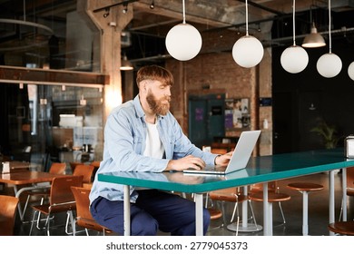 Freelancer with beard using notebook computer at high table in loft style workspace, suited for technology, entrepreneurship, online business, and work anywhere themes