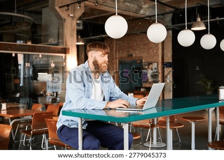 Freelancer with beard using notebook computer at high table in loft style workspace, suited for technology, entrepreneurship, online business, and work anywhere themes