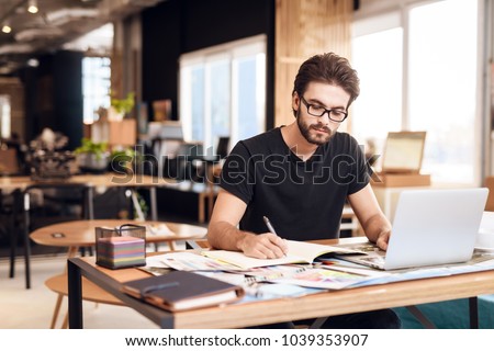 Freelancer bearded man in t-shirt taking notes at laptop sitting at desk.