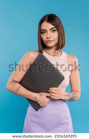 freelancer, brunette young woman with short hair, tattoos and nose piercing holding laptop on blue background, generation z, summer trends, attractive, remote work, everyday style