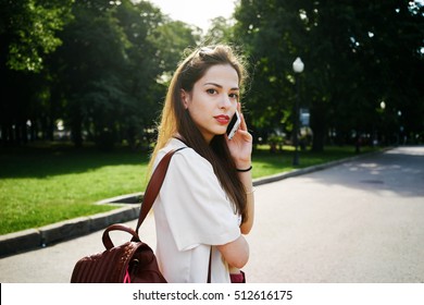 A Freelancer Is Calling To A Client By A Mobile Phone To Make An Appointment. Young Brunette Woman Is Holding A Smart-phone While Having Rest In A City Park.