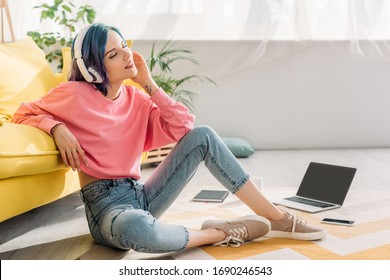 Freelancer With Colorful Hair, Closed Eyes And Headphones Listening Music Near Sofa, Laptop And Smartphone On Floor